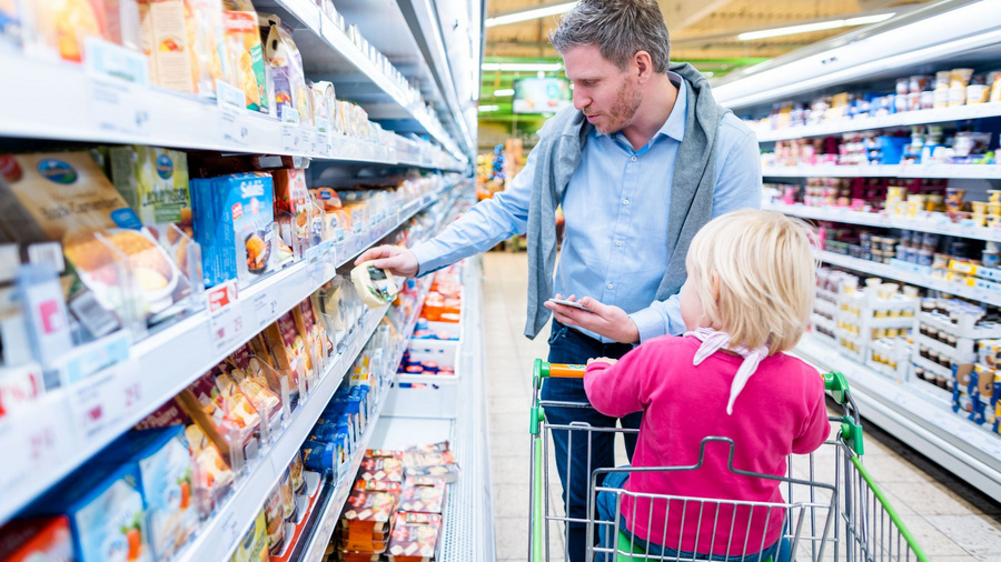 Mann mit Kind, das in einem Einkaufswagen sitzt, vor einem Kühlregal im Supermarkt