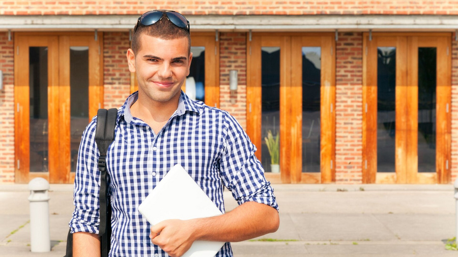 Student mit Laptop in der Hand steht vor Uni.